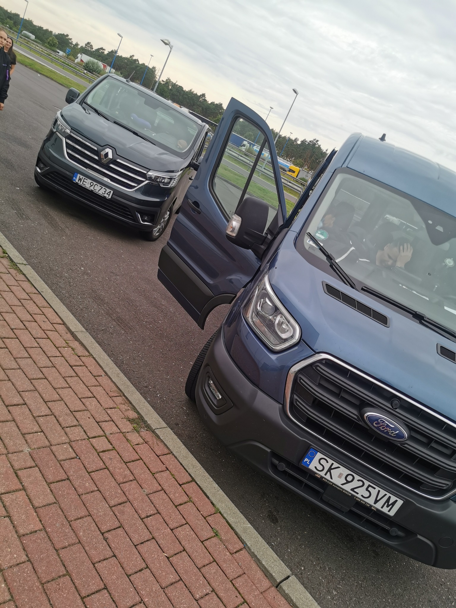 Dwa busy, Renault Trafic i Ford Transit, zaparkowane obok siebie na parkingu, w Fordzie Transit otwarte drzwi kierowcy, widoczny kierowca.