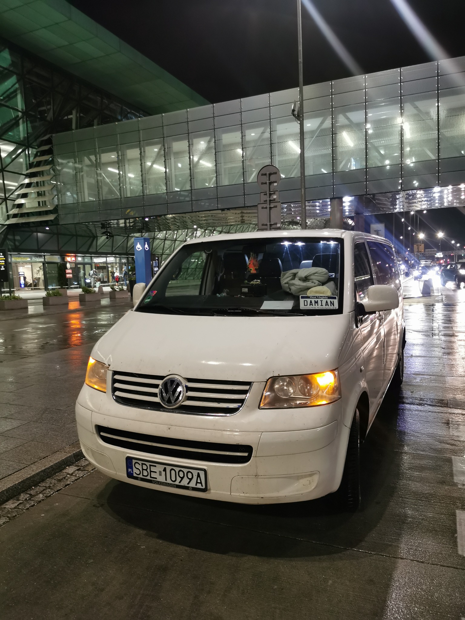 Biały bus Volkswagen parkuje nocą przed nowoczesnym terminalem lotniska, z kartką 'DAMIAN' na desce rozdzielczej.