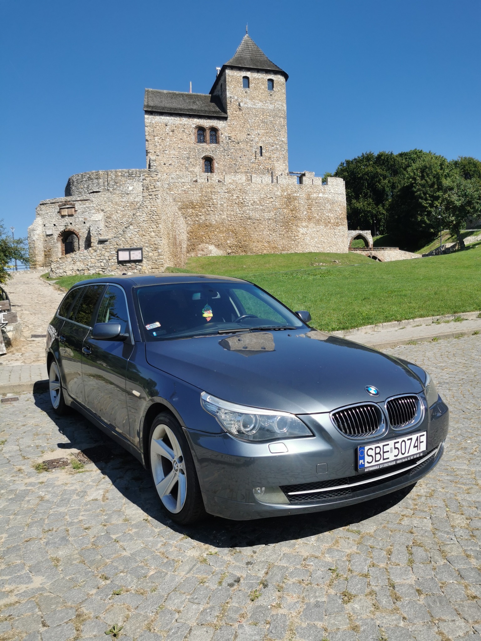 Szary samochód typu kombi marki BMW zaparkowany na brukowanej drodze z widokiem na średniowieczny kamienny zamek z wieżą na wzgórzu w słoneczny dzień.