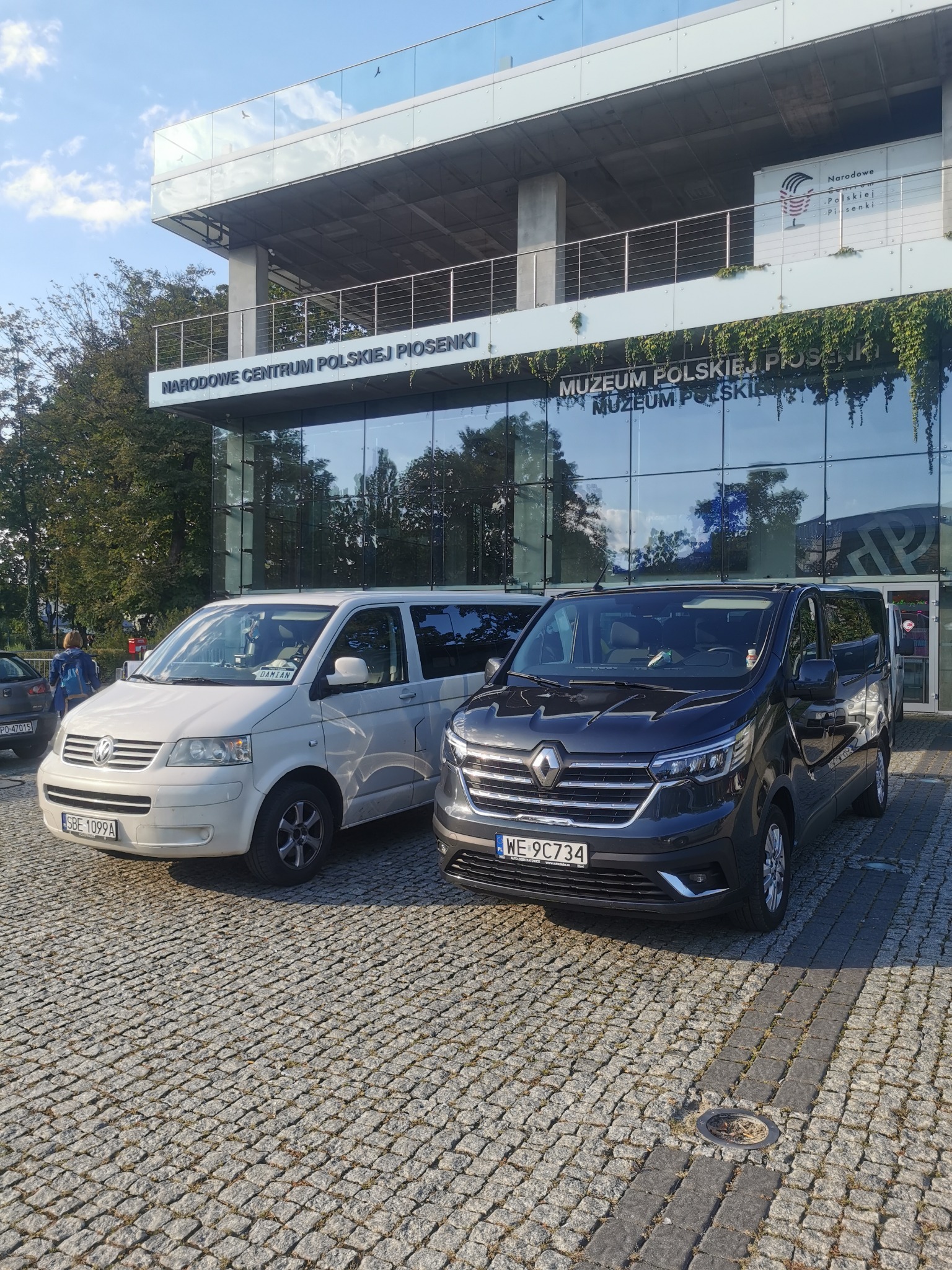 Dwa busy, biały Volkswagen i ciemny Renault, zaparkowane przed nowoczesnym budynkiem Narodowego Centrum Polskiej Piosenki w Opolu, widok z chodnika z kostki brukowej.