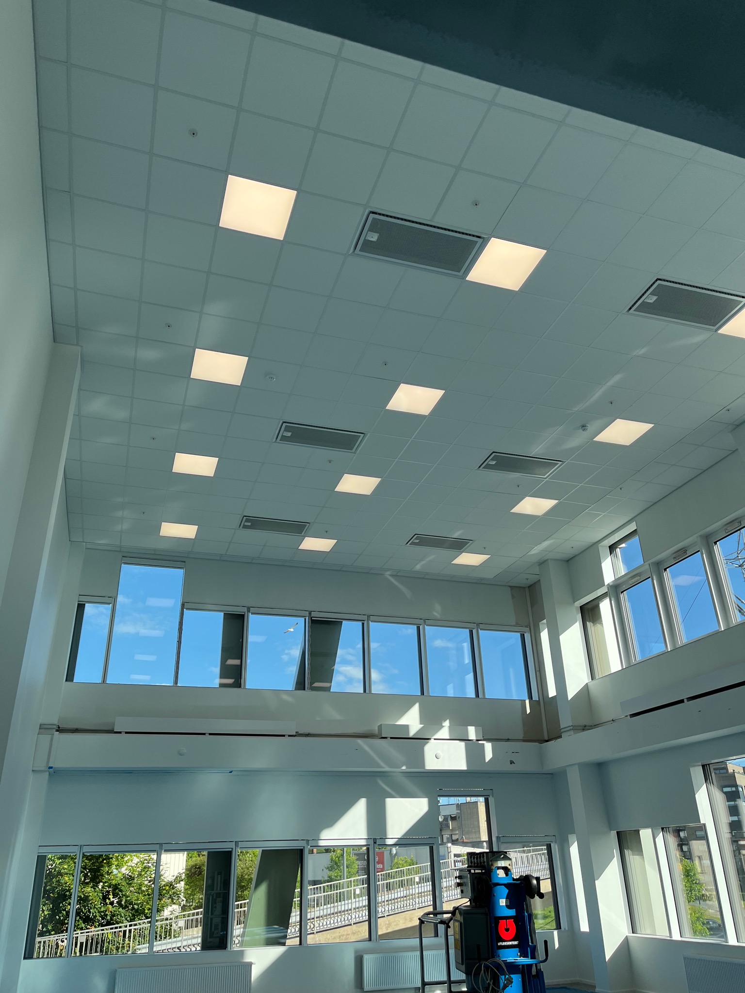 Wnętrze biurowe z widocznym sufitem kasetonowym z wbudowanym oświetleniem i systemem wentylacji, duże okna z widokiem na zewnątrz, maszyna czyszcząca podłogi w rogu pomieszczenia.