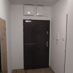 Wnętrze przedpokoju z drewnianą podłogą, ciemnymi drzwiami wejściowymi i lustrem z wieszakami po lewej stronie. Na suficie widoczne okrągłe oświetlenie oraz dwa białe panele wentylacyjne nad drzwiami.