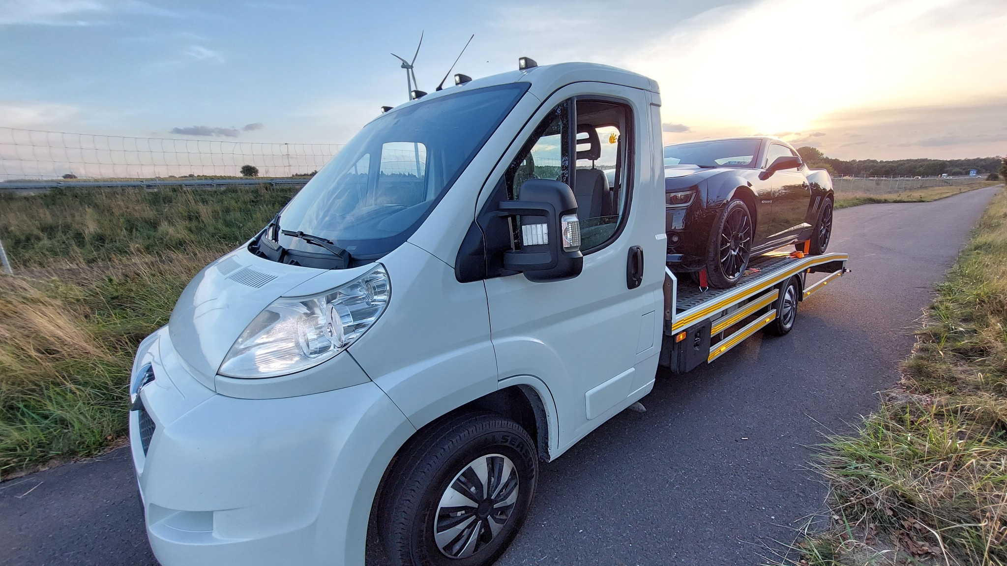 Biała laweta z czarnym sportowym samochodem na platformie, widok z boku na tle zachodzącego słońca i turbin wiatrowych.
