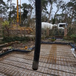 Domi-Rem - Wylewanie betonu na zbrojenie fundamentu domu jednorodzinnego. W tle widoczna betoniarka i pompa do betonu. Prace budowlane w toku, teren częściowo zadrzewiony.