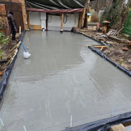 Domi-Rem - Wylana, świeża betonowa posadzka na zewnątrz budynku z widocznymi deskami szalunkowymi i folią. W tle budynek z cegły i zadaszone wejście.