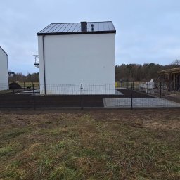 Domi-Rem - Nowoczesny dom z białymi ścianami i ciemnym dachem, otoczony czarnym ogrodzeniem panelowym. W tle widoczny budynek w trakcie budowy.