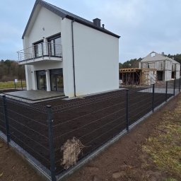 Domi-Rem - Nowoczesny dom z szarym ogrodzeniem panelowym na świeżo przygotowanym gruncie. W tle dom w trakcie budowy i las.