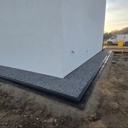 Domi-Rem - Wykończona opaska z kostki brukowej i grysu wokół narożnika białego budynku, widok z poziomu gruntu, z fragmentem ogrodzenia panelowego w tle.