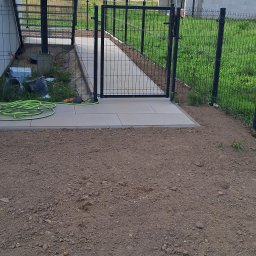Domi-Rem - Nowoczesne ogrodzenie panelowe z bramką, prowadzące do betonowego chodnika. W tle widoczna zieleń trawy i fragment budynku. Na pierwszym planie ziemia.