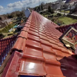 Nowo położona, błyszcząca, czerwona dachówka ceramiczna z widocznym oknem dachowym i elementami wykończeniowymi, na tle błękitnego nieba i zabudowań.