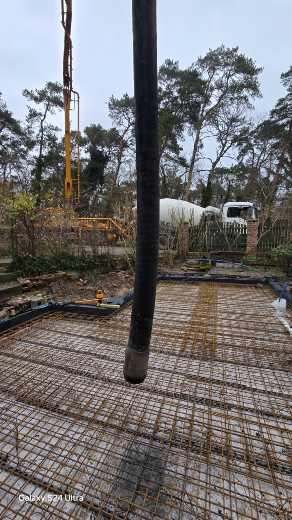 Wylewanie betonu na zbrojenie fundamentu domu jednorodzinnego. W tle widoczna betoniarka i pompa do betonu. Prace budowlane w toku, teren częściowo zadrzewiony.