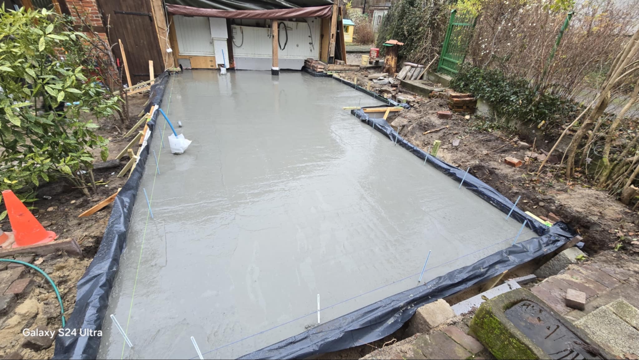 Świeżo wylana, gładka betonowa wylewka na zewnątrz, zabezpieczona czarną folią i drewnianymi deskami. W tle widoczny budynek gospodarczy i zieleń.