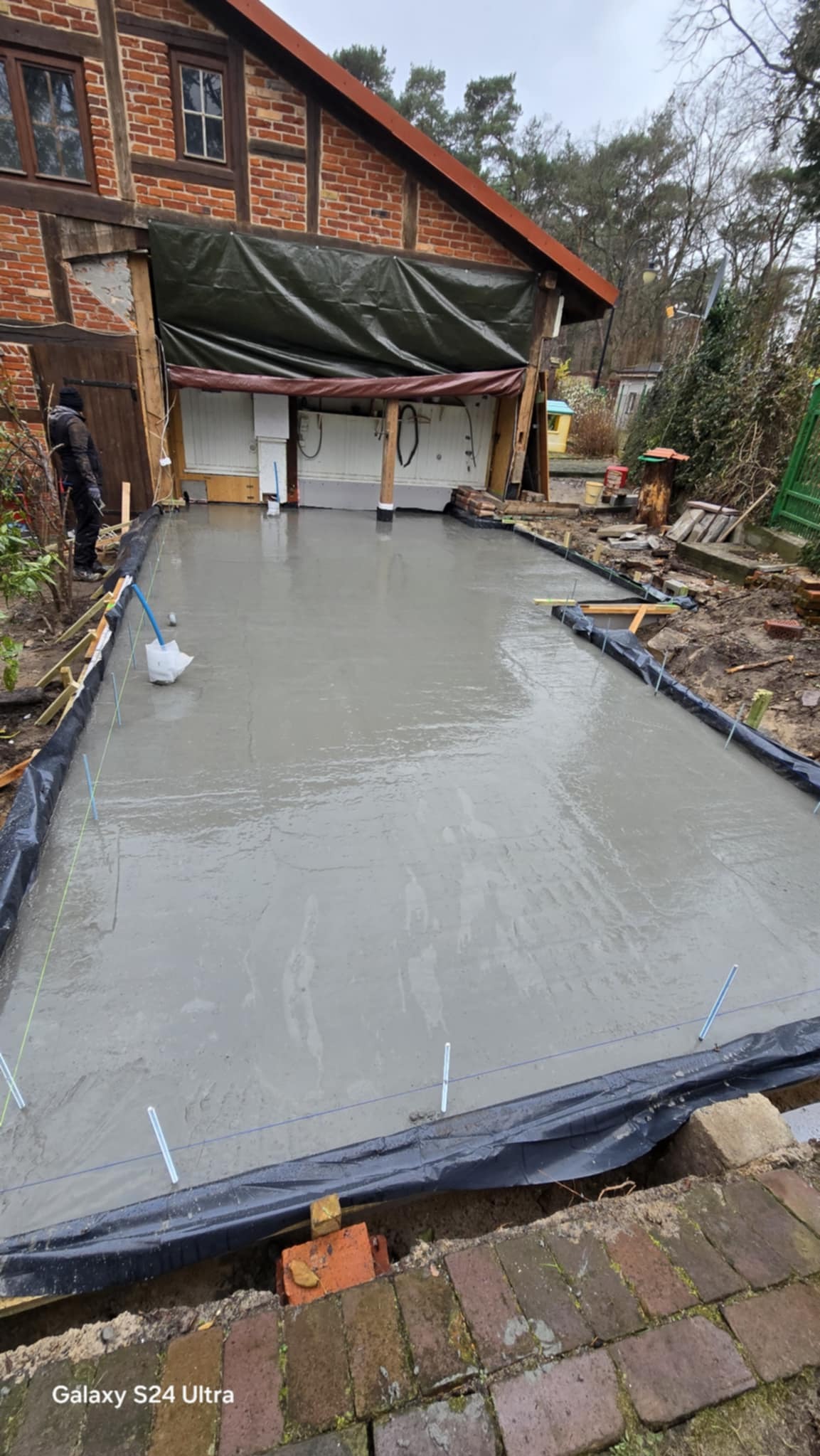 Wylana, świeża betonowa posadzka na zewnątrz budynku z widocznymi deskami szalunkowymi i folią. W tle budynek z cegły i zadaszone wejście.