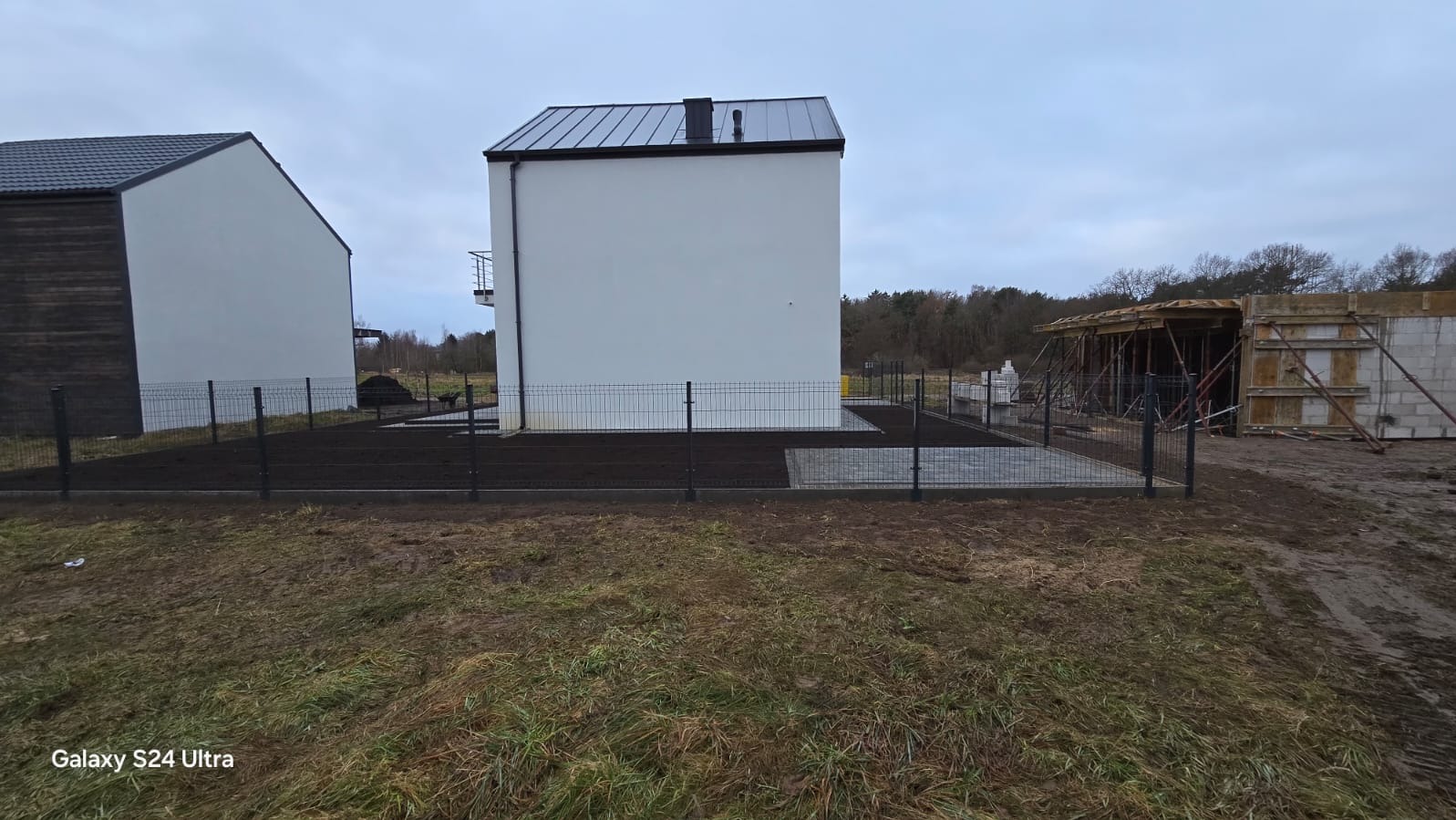 Nowoczesny dom z białymi ścianami i ciemnym dachem, otoczony czarnym ogrodzeniem panelowym. W tle widoczny budynek w trakcie budowy.