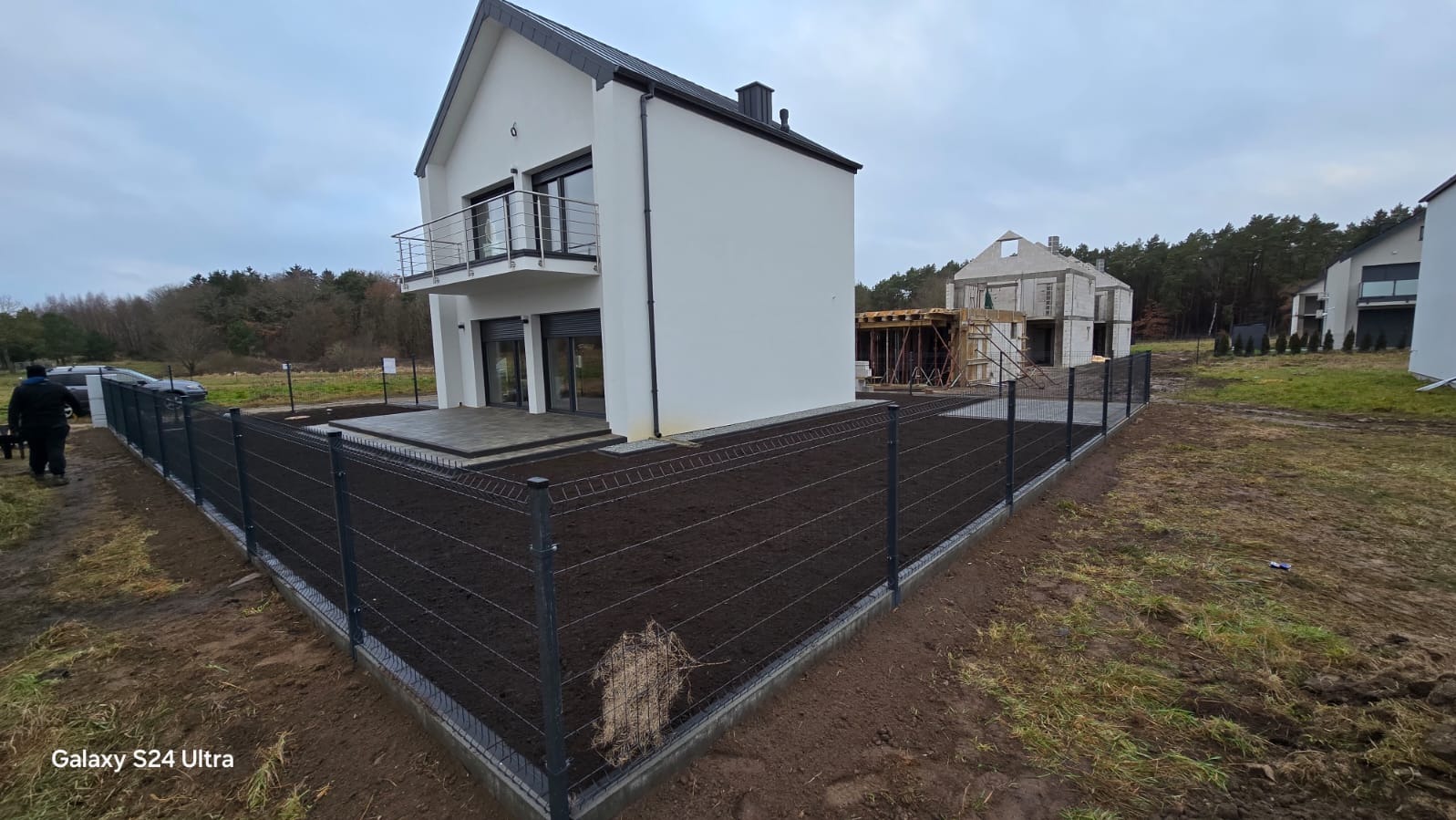 Nowoczesny dom z szarym ogrodzeniem panelowym na świeżo przygotowanym gruncie. W tle dom w trakcie budowy i las.