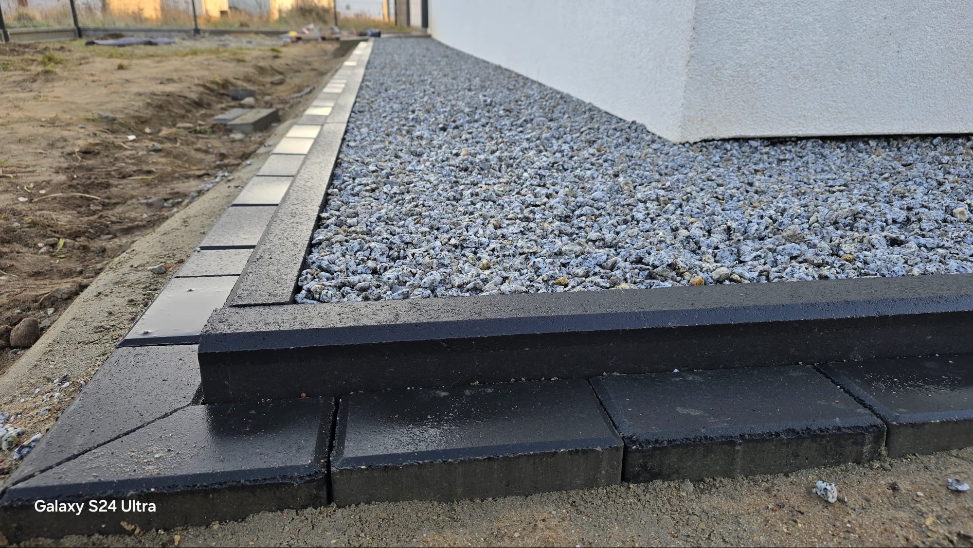 Ułożona, czarna obwódka z kostki brukowej przy białej ścianie budynku, wypełniona szarym żwirem. Widoczne ślady wilgoci na kostce.