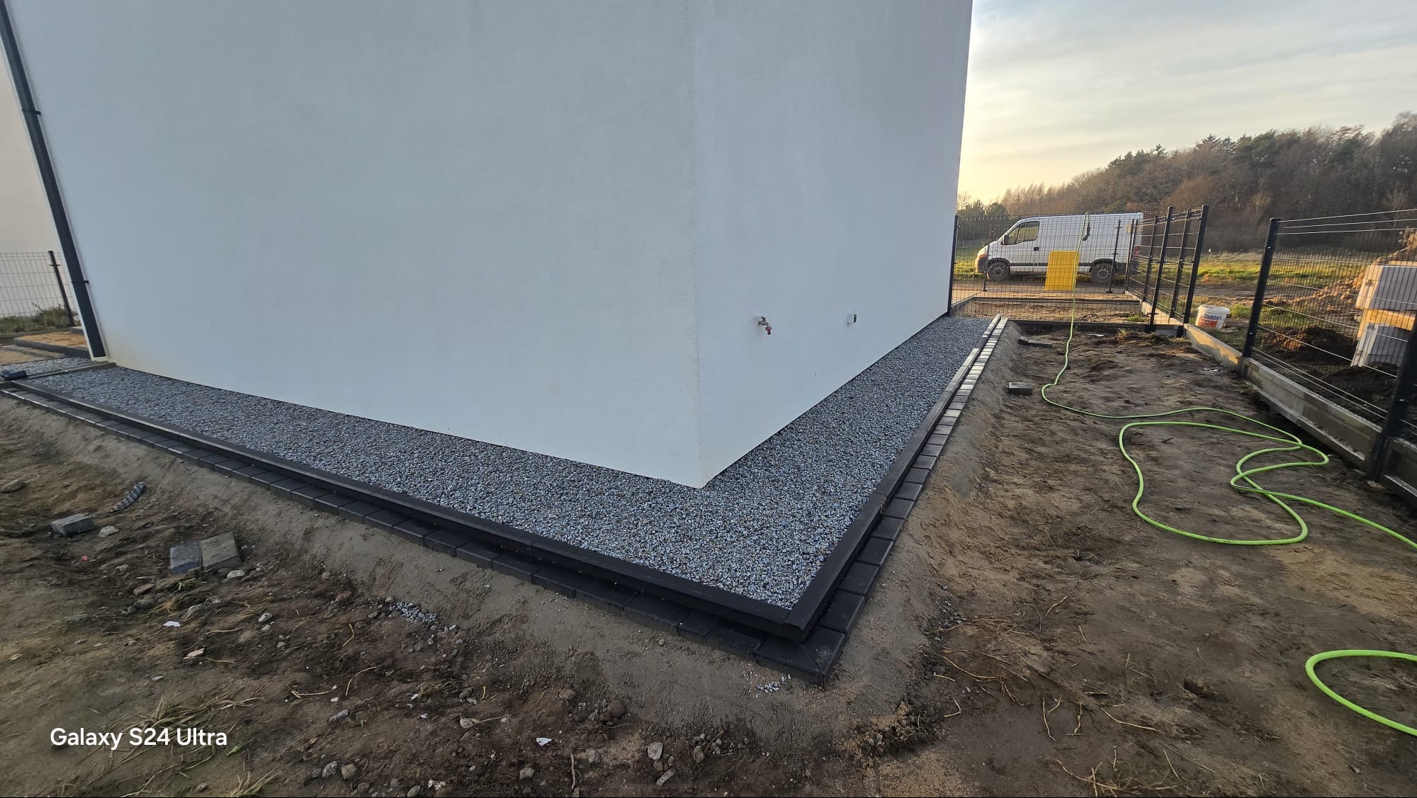 Wykończona opaska z kostki brukowej i grysu wokół narożnika białego budynku, widok z poziomu gruntu, z fragmentem ogrodzenia panelowego w tle.