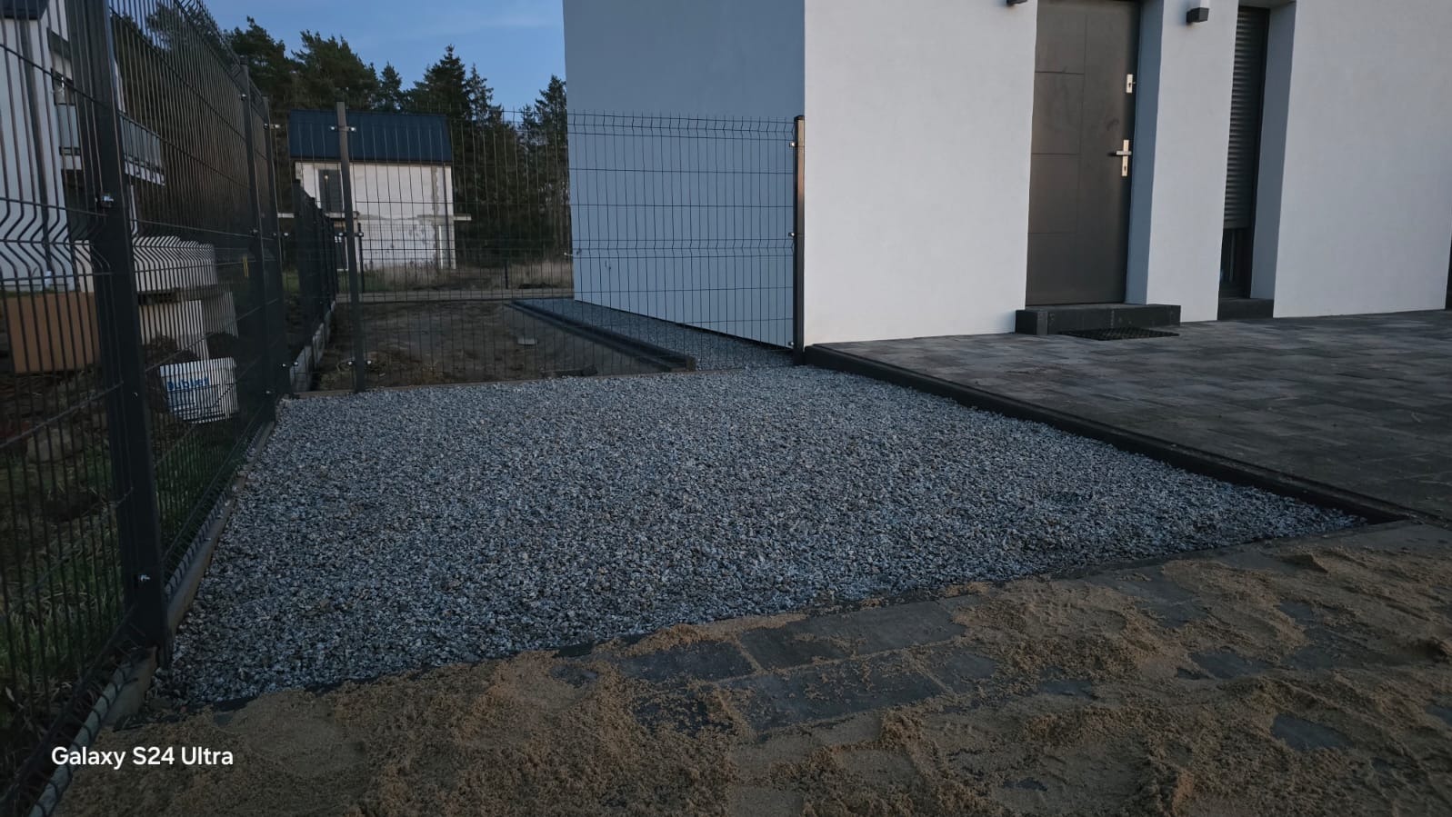 Nowoczesny dom z szarą elewacją, ogrodzeniem panelowym i wysypanym żwirem podjazdem. Widoczne kostki brukowe i piasek. Ujęcie z poziomu gruntu.