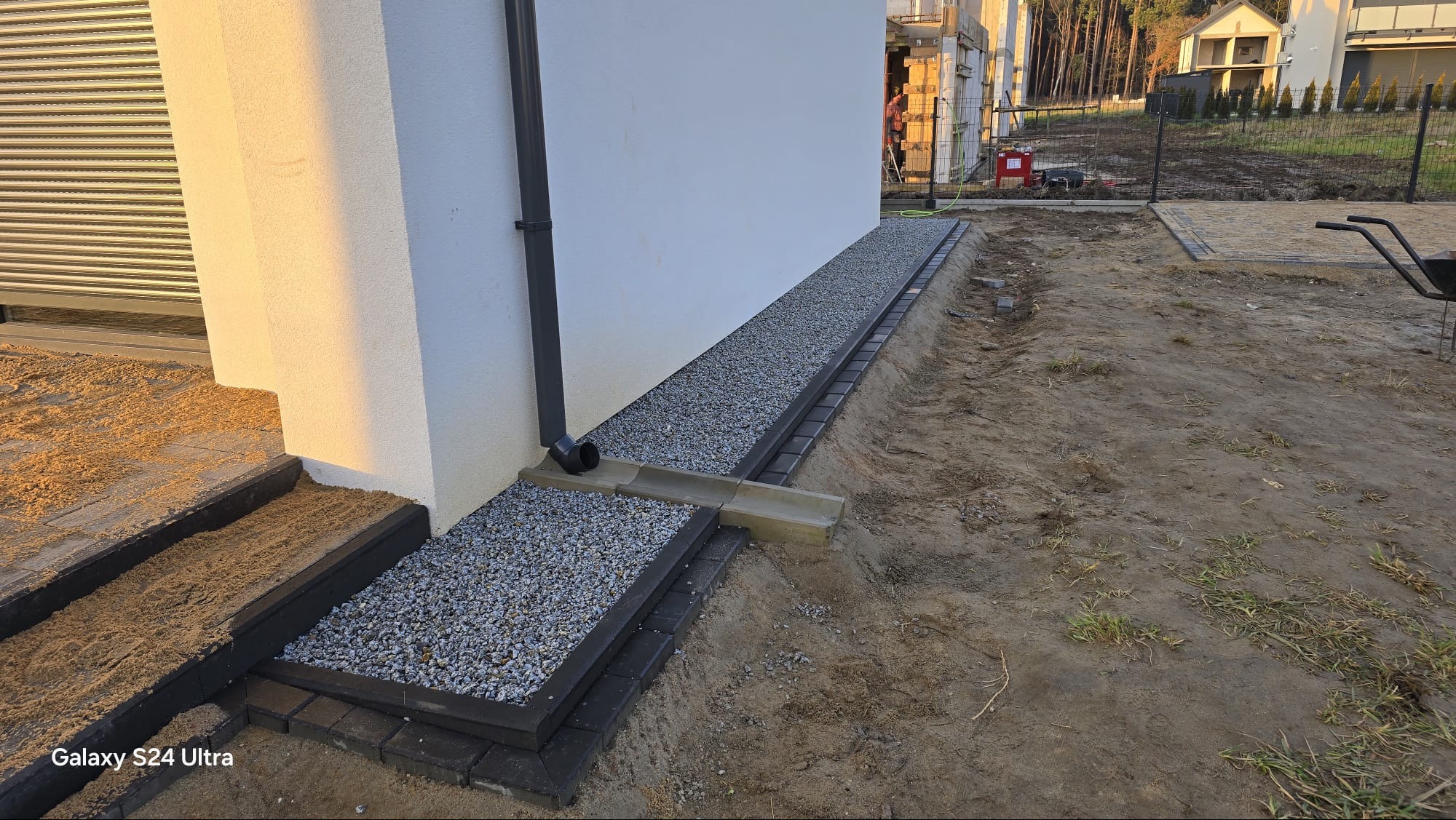 Wykończona opaska drenażowa z kostki brukowej i grysu przy ścianie domu z rurą spustową. Widoczne schody wejściowe z kostki i teren wokół w trakcie prac ziemnych.