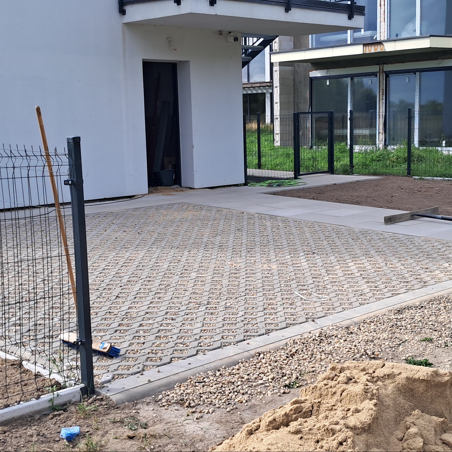 Ułożona ażurowa nawierzchnia z kostki betonowej na podjeździe, widoczny fragment ogrodzenia panelowego i sterta piasku. Prace wykończeniowe na posesji.