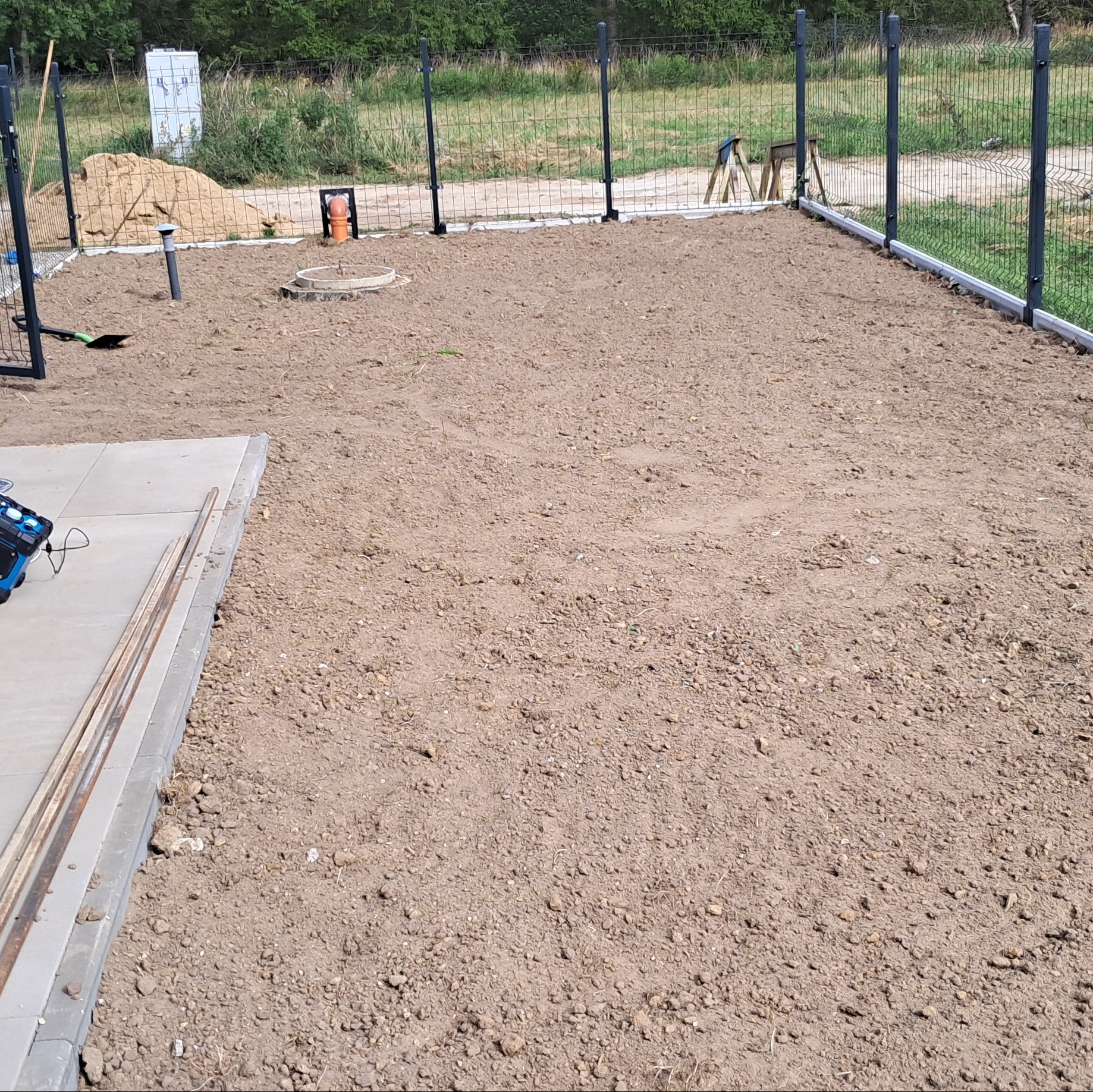 Przygotowany teren pod trawnik z widocznym ogrodzeniem panelowym i elementami instalacji. Widoczne narzędzia i materiały budowlane.