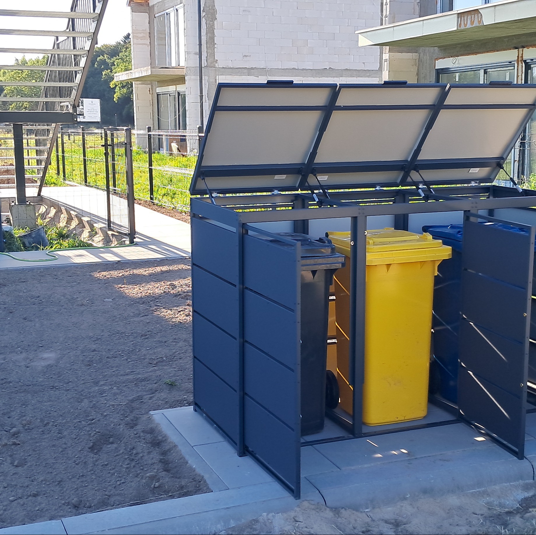 Metalowa zabudowa śmietnikowa z otwartymi klapami, wewnątrz kolorowe pojemniki na odpady, na tle surowego budynku i ogrodzenia panelowego.