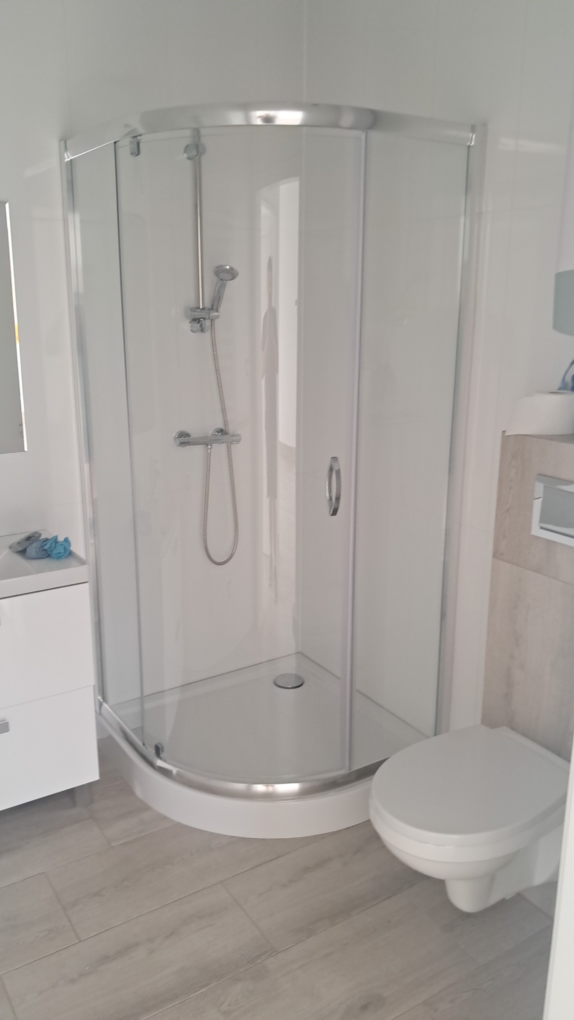 Nowoczesna kabina prysznicowa z chromowanymi detalami w jasnej łazience z drewnopodobną podłogą i wiszącą toaletą. Minimalistyczny design.