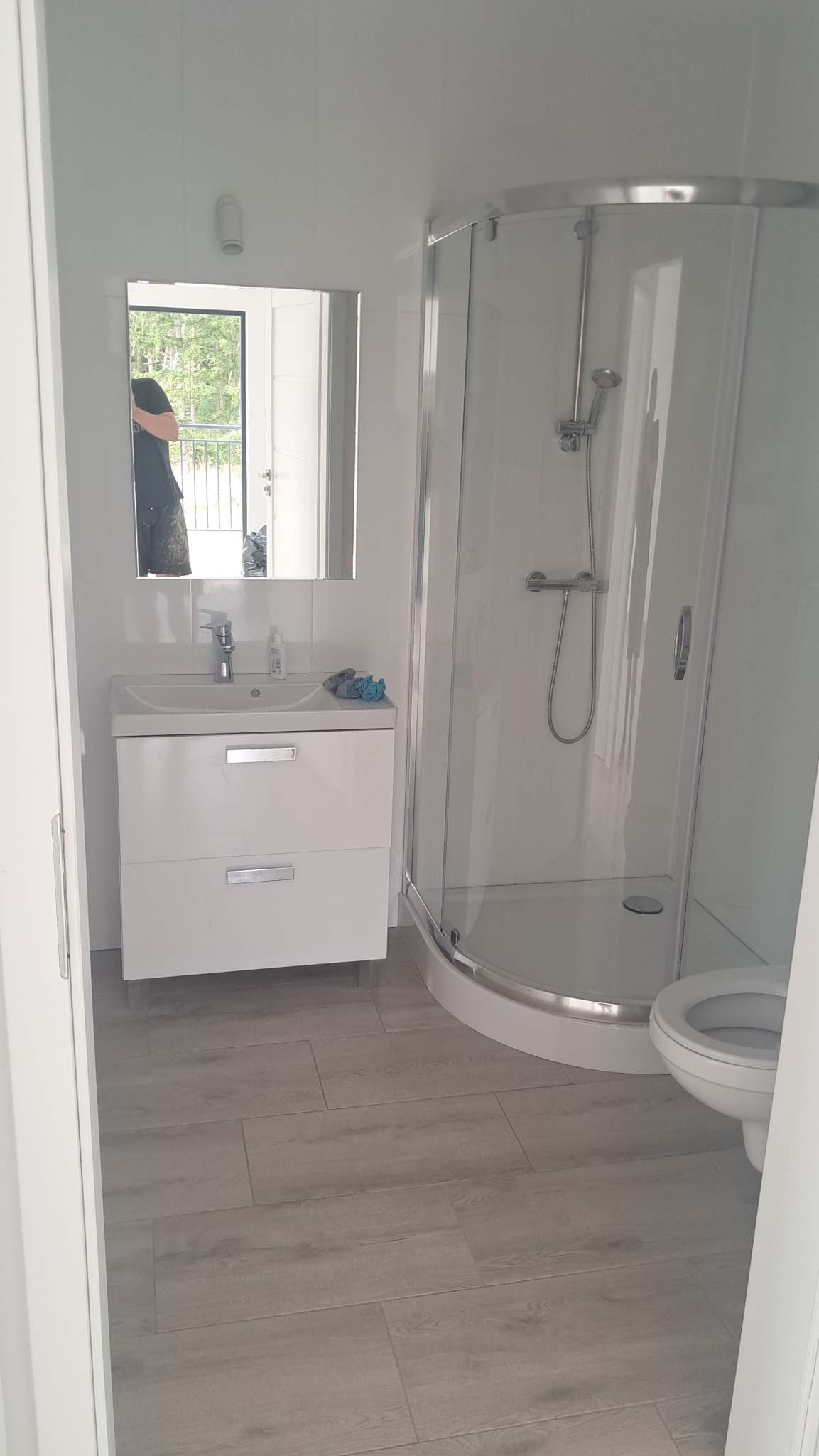 Jasna łazienka po remoncie: kabina prysznicowa, umywalka z szafką i toaleta. Na podłodze panele imitujące drewno. W lustrze odbija się fragment balkonu.