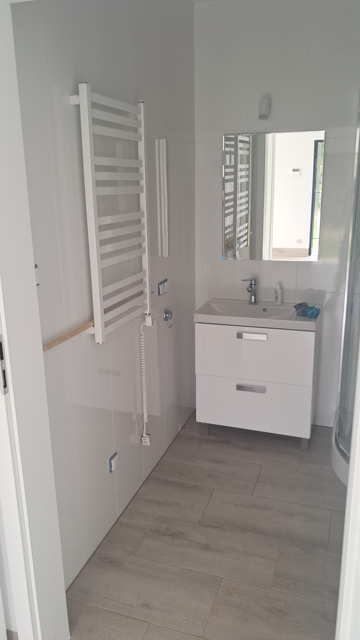Biała łazienka z grzejnikiem drabinkowym, szafką z umywalką i lustrem. Widoczne płytki na ścianach i podłodze imitujące drewno. Częściowo zamontowany grzejnik.