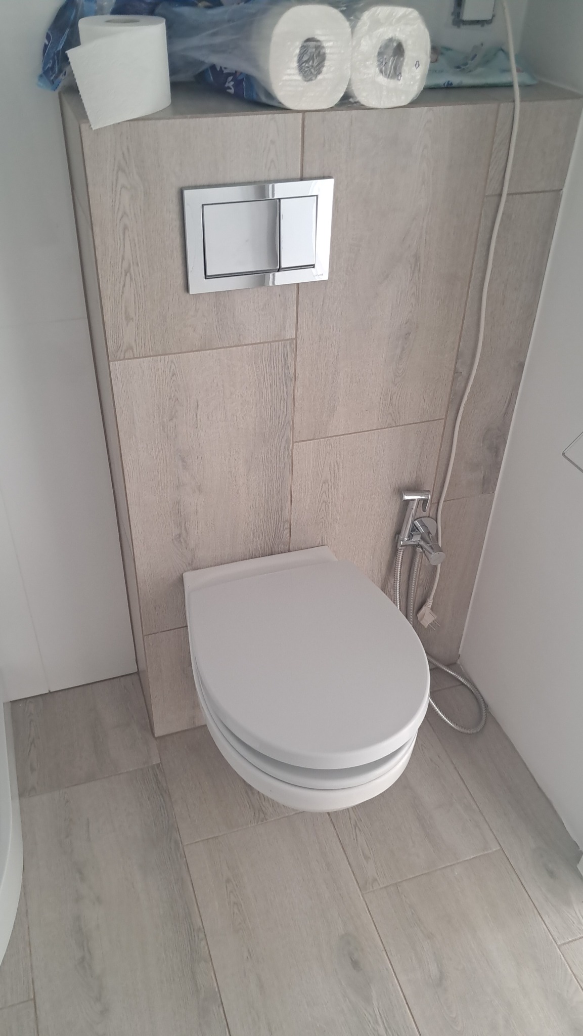 Nowoczesna toaleta z bidetką i przyciskiem spłukującym, wykończona płytkami imitującymi drewno. Widoczne rolki papieru toaletowego na obudowie stelaża.