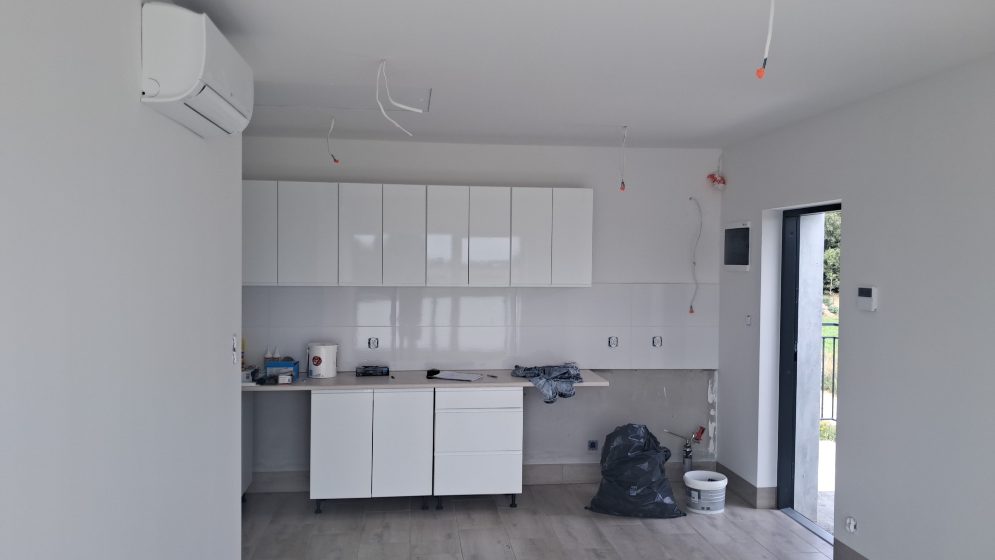 Wnętrze w trakcie wykańczania: białe szafki kuchenne, płytki na ścianie, widoczne przewody elektryczne, podłoga z paneli, klimatyzator na ścianie, ślady prac remontowych.