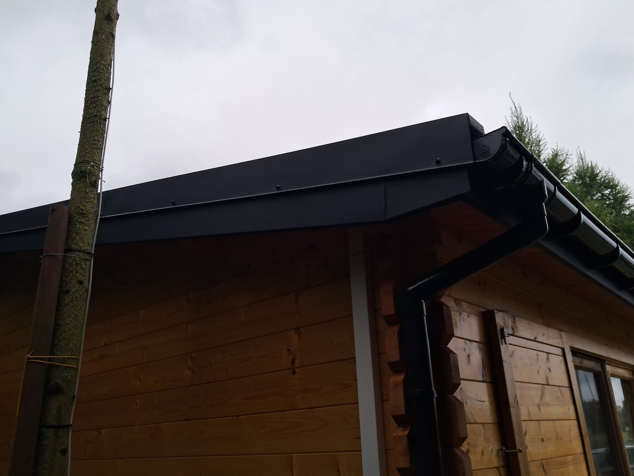 Fragment drewnianej altany z czarnym dachem i systemem rynnowym, widoczne elementy konstrukcyjne i orynnowanie z bliska.