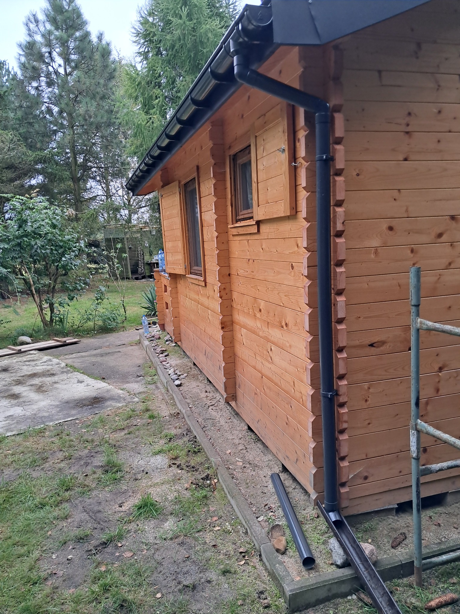 Drewniana altana z zamontowanym systemem rynnowym w kolorze grafitowym, widoczne elementy konstrukcyjne z bali i fragment drabiny.