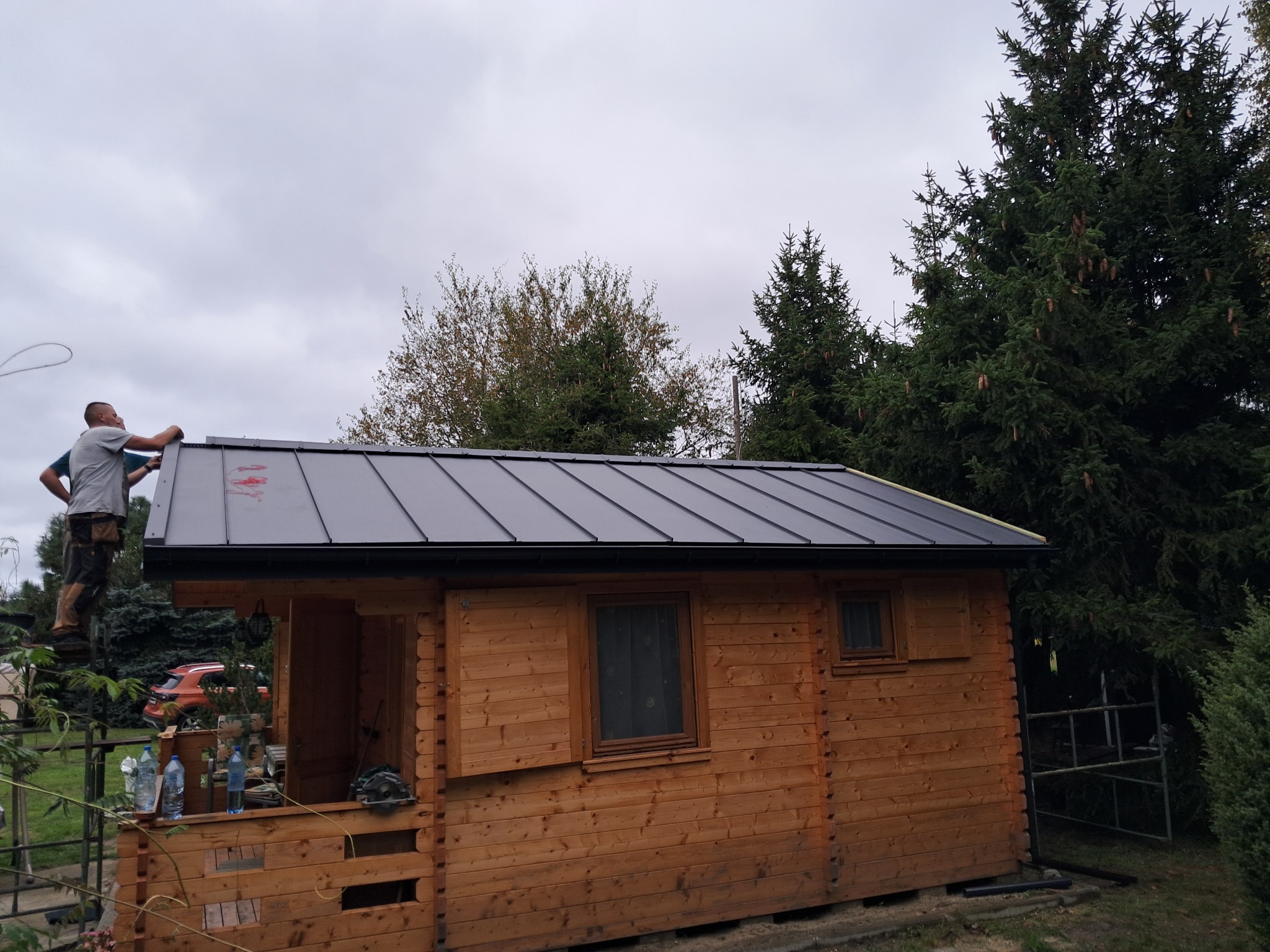 Montaż czarnego dachu z blachy na drewnianej altanie z oknem, widoczni dwaj pracownicy na dachu, w tle drzewa iglaste i samochód