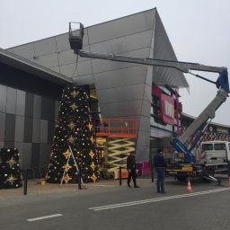 F.H.U. Top-Marek Marek Bis - Montaż świątecznej dekoracji na zewnątrz budynku w Pysznicy przy użyciu podnośnika koszowego, widoczna choinka ozdobiona gwiazdami i bombkami.