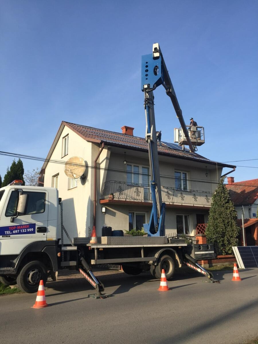 Montaż paneli słonecznych na dachu domu jednorodzinnego z użyciem podnośnika koszowego, widoczna ekipa pracująca na wysokości, panele przygotowane do montażu obok samochodu, zabezpieczenie terenu...