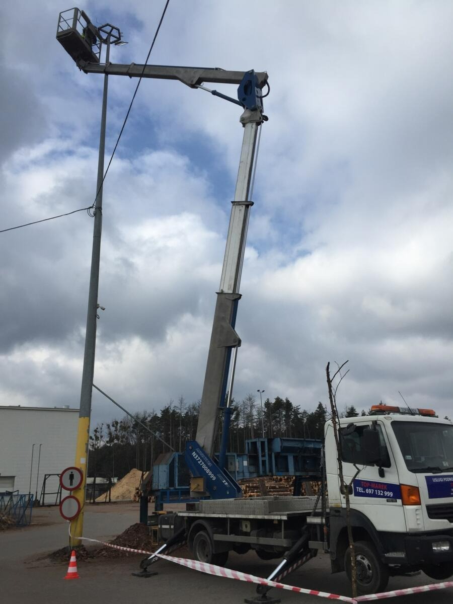 Wysięgnik koszowy na pojeździe ustawiony przy słupie oświetleniowym, widoczne znaki drogowe i pomarańczowy pachołek, w tle budynki i drzewa.