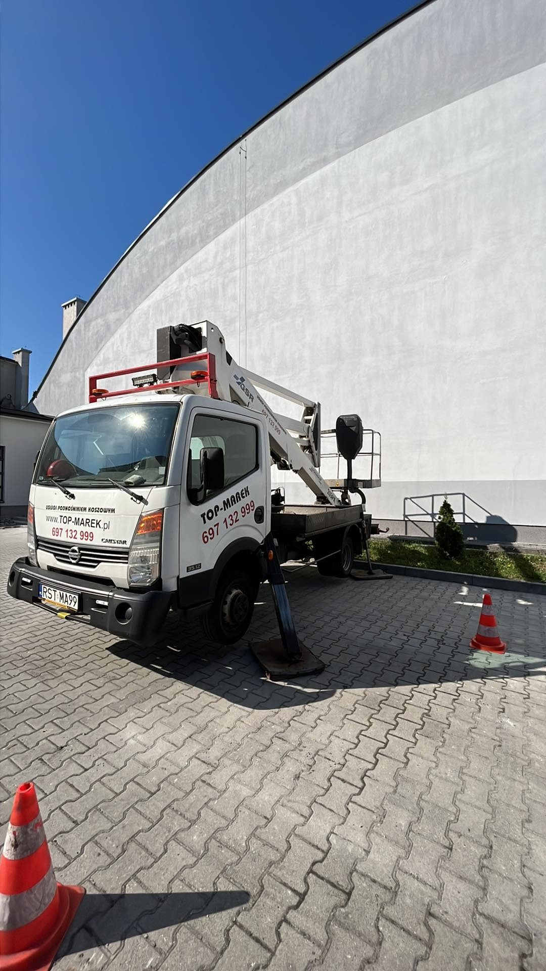 Podnośnik koszowy marki GSR na białym samochodzie dostawczym z rozłożonymi podporami, zaparkowany na kostce brukowej przed wysoką, jasną ścianą budynku, z dwoma pachołkami ostrzegawczymi w pobliżu.