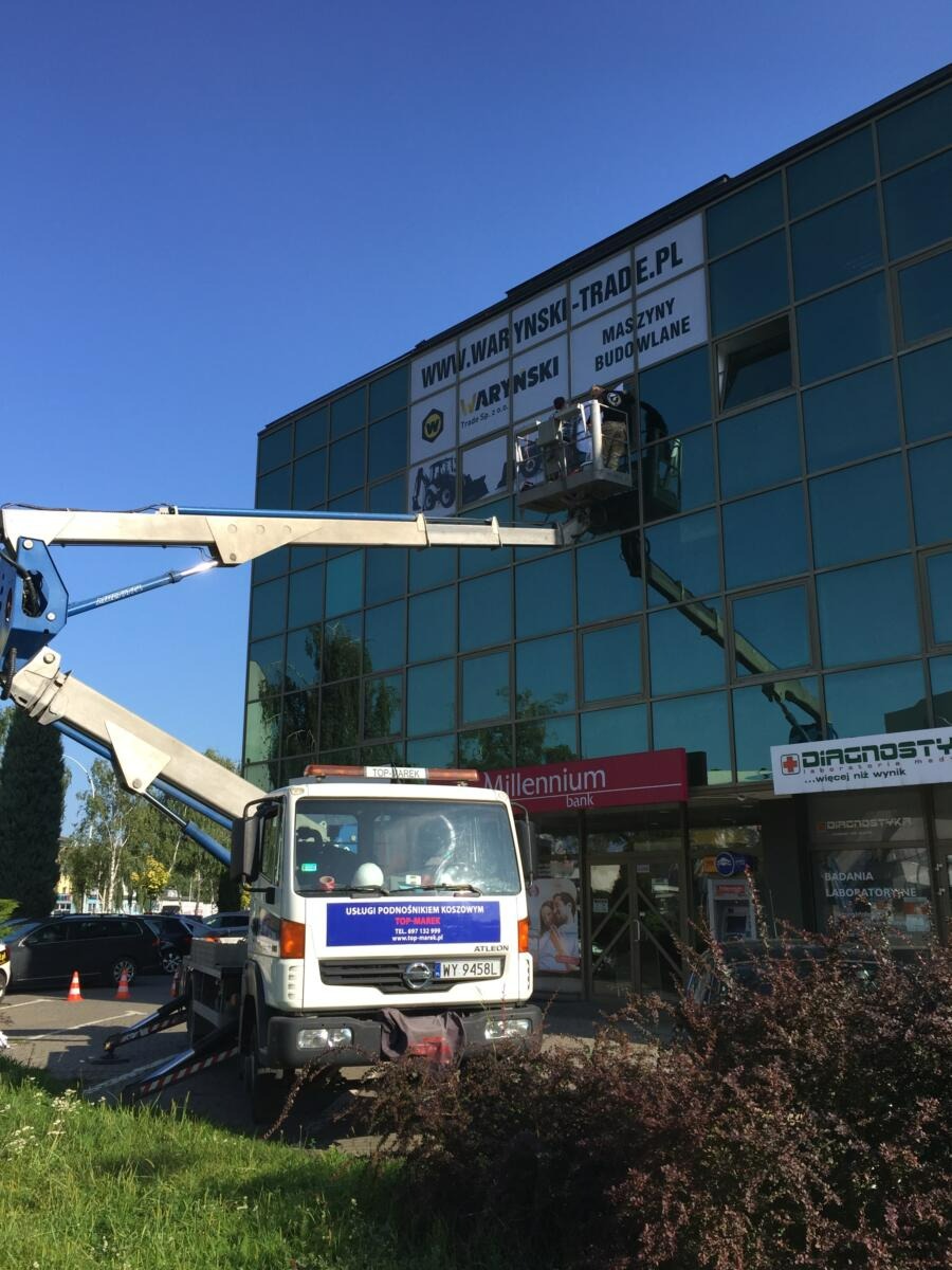 Montaż baneru reklamowego na szklanym budynku biurowym z użyciem podnośnika koszowego; widoczny fragment logotypu firmy Millennium Bank