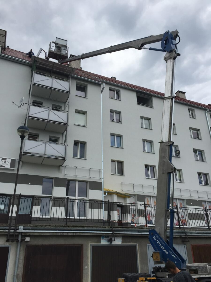 Praca z podnośnika koszowego na dachu budynku mieszkalnego, widoczne balkony i elementy elewacji, pochmurne niebo w tle.