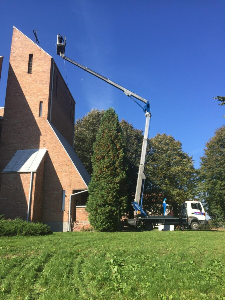 Prace wysokościowe przy kościele: podnośnik koszowy z pracownikiem przy krzyżu na szczycie wieży, ceglana elewacja, błękitne niebo.