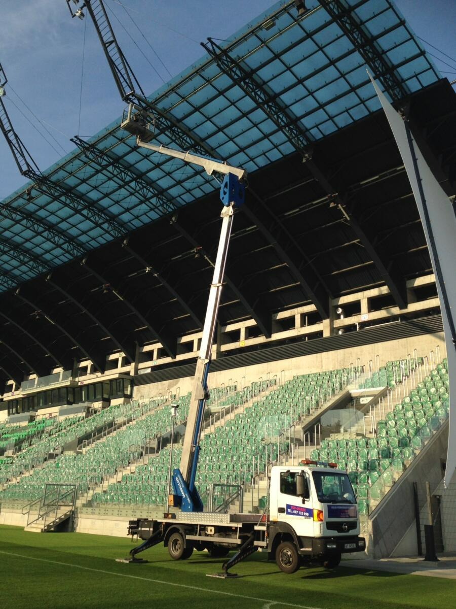 Podnośnik koszowy na stadionie, konserwacja dachu z pleksi, widoczne trybuny i murawa.