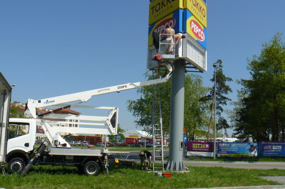 Montaż reklamy zewnętrznej na wysokim słupie przez dwóch pracowników z podnośnika koszowego na tle błękitnego nieba i zieleni