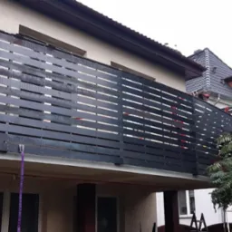 Nowoczesna balustrada balkonowa z poziomych, ciemnych desek, widoczna z dołu, z elementami mocującymi i zielenią w tle.