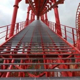 Stalowe schody z ażurowymi stopniami prowadzące w górę, czerwona konstrukcja nośna z rur i belek, widok z dołu w kierunku nieba.