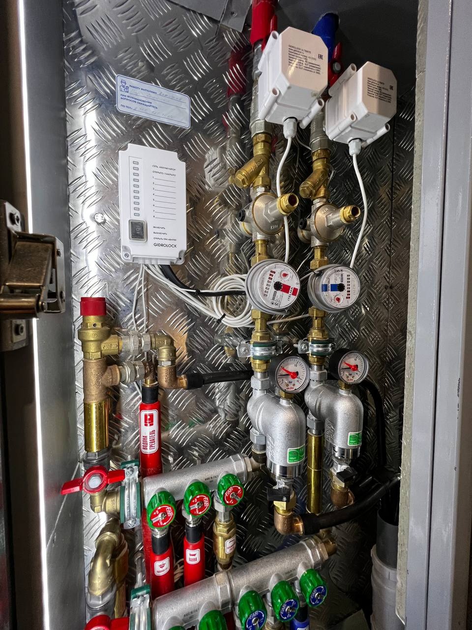 Węzeł wodomierzowy z automatycznym systemem odcinającym dopływ wody, zamontowany w metalowej szafce z blachy ryflowanej.