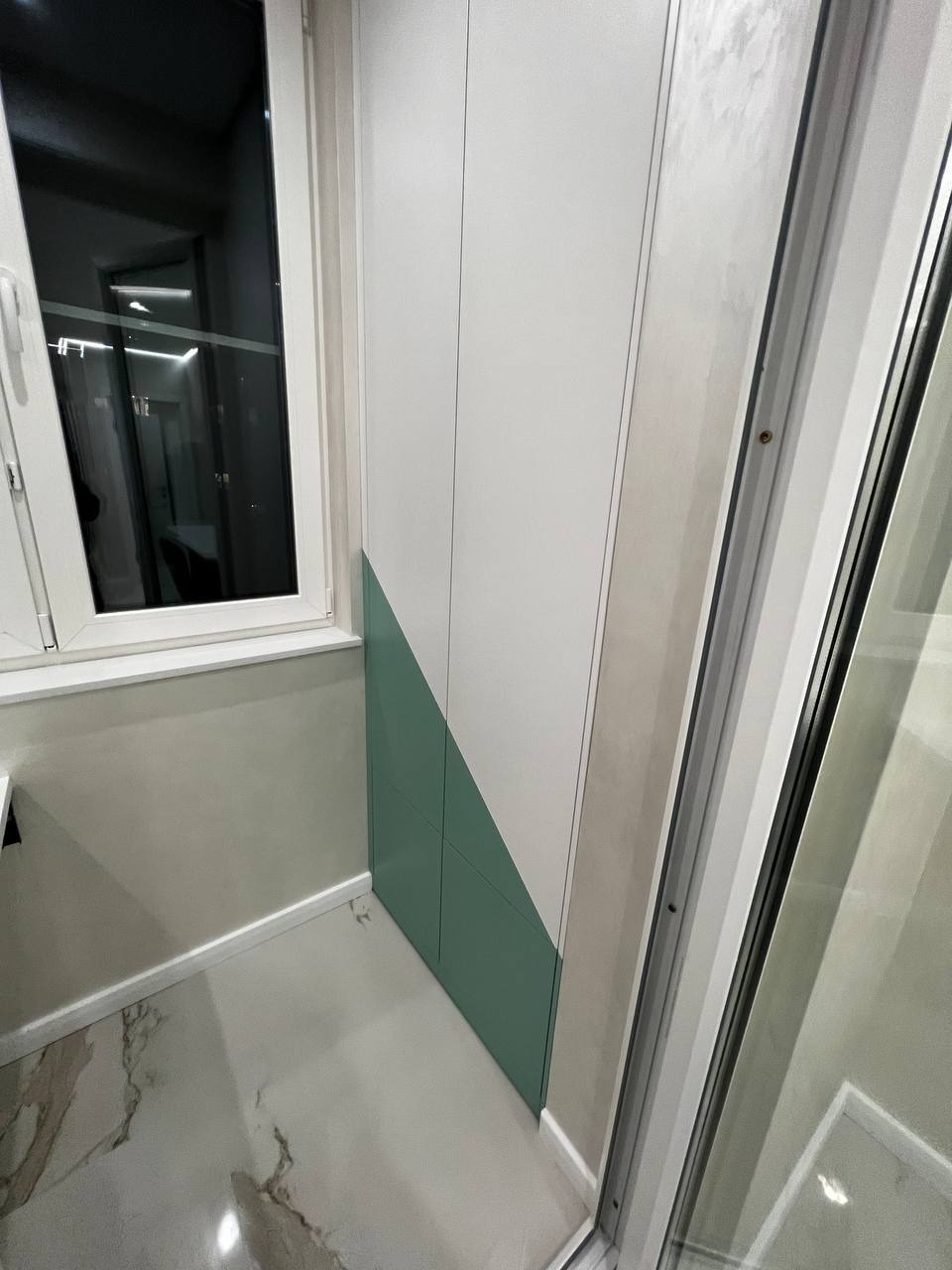 Wnętrze balkonu po remoncie z zabudową szafy w kolorach białym i zielonym, widoczne marmurowe płytki podłogowe i okno z widokiem na zewnątrz.