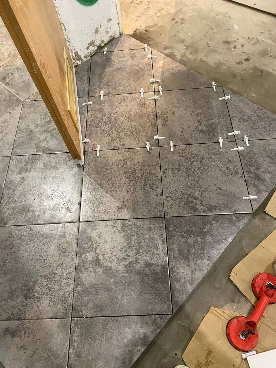 Ułożone płytki podłogowe w szarym odcieniu z krzyżykami dystansowymi i przyssawką do płytek, widoczne przy drzwiach w trakcie prac wykończeniowych.