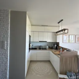 Nowoczesna, jasna kuchnia z białymi szafkami, drewnianym blatem i okrągłym dywanem w stylu boho; fragment ściany z białej cegły dekoracyjnej.