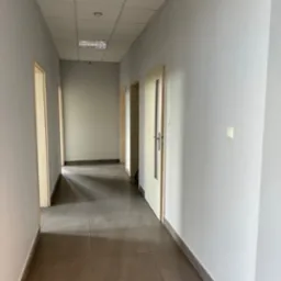 Pomalowany na szaro korytarz biurowy z brązowymi płytkami podłogowymi i białymi drzwiami, widok na sufit z oświetleniem rastrowym.